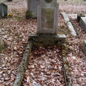 obmurowany grób i wysoka płyta nagrobna z płaskorzeźbioną głową Chrystusa i tablicą epitafijną; za nim inne nagrobki i pnie drzew