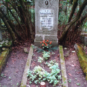 obmurowany grób z zielenią wewnątrz oraz czerwonym zniczem i wysoka stela z napisami, medalionem portretowym i krzyżem łacińskim