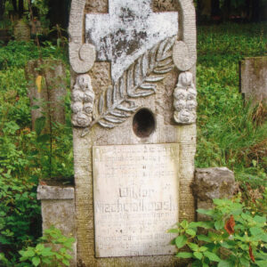 zabrudzona, wysoka płyta nagrobna z tablicą epitafijną, w górnej części krzyż grecki z ukośną gałązką palmową; za nagrobkiem teren cmentarza i drzewa