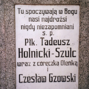 biała, prostokątna tablica z czarnymi napisami wtopiona w płytę nagrobną