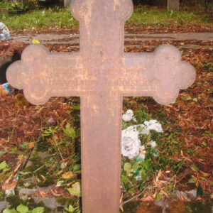 pordzewiały krzyż łaciński o trójlistnie zakończonych ramionach z napisami, za nim teren cmentarza, inne nagrobki i drzewa