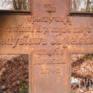 napisy wyryte na przecięciu ramion pordzewiałego krzyża, za krzyżem teren cmentarza