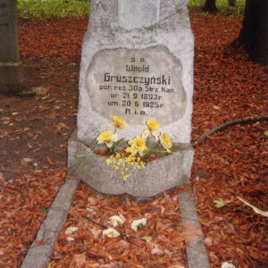 obmurowany grób i obelisk z tablicą epitafijną ozdobiony żółtymi, sztucznymi kwiatami; za nim inne nagrobki i pnie drzew, teren pokryty suchymi liśćmi