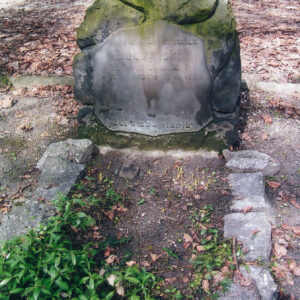 grób otoczony kamieniami i omszona stela w formie głazu z tablicą epitafijną; z tyłu inne nagrobki i teren cmentarza