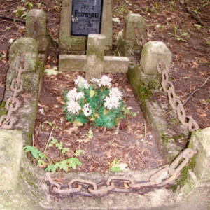 grób obramowany murkiem z kamiennymi słupkami, które są połączone łańcuchami i stela z tablicą epitafijną; z tyłu teren cmentarza