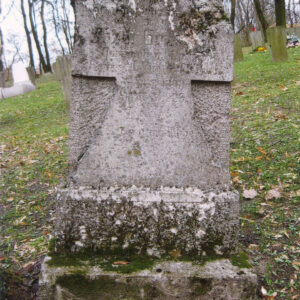 zabrudzony i uszkodzony nagrobek w formie prostokątnej płyty z krzyżem, którego dolna belka rozszerza się ku cokołowi; z tyłu inne nagrobki i pnie drzew