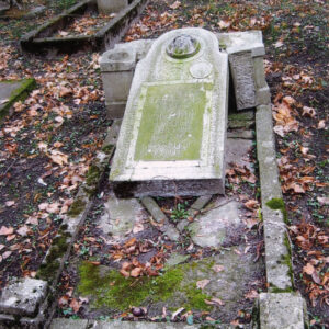 zniszczony obmurowany grób, oddzielona od cokołu wysoka stela leżąca wewnątrz obramowania; wokół teren cmentarza z innymi nagrobkami