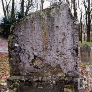 znacznie uszkodzona tablica z licznymi obtłuczeniami i nieczytelną inskrypcją, za nią teren cmentarza z innymi nagrobkami i drzewami