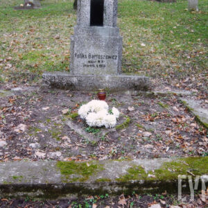 szerokie obmurowanie grobu pokryte mchem, pośrodku białe kwiaty i znicz, za fragmentem steli teren cmentarza