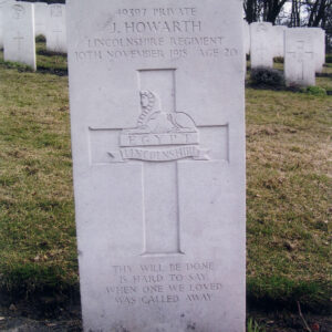 Biała płyta nagrobna z napisami i znakiem pułkowym pośrodku krzyża łacińskiego.  Za nią inne nagrobki i drzewa.