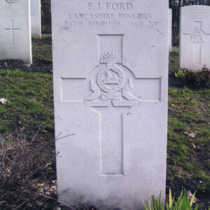 biała, pionowa płyta nagrobna z napisami i wyżłobionym krzyżem łacińskim, wewnątrz którego wyryto znak pułku, za nią inne nagrobki i drzewa