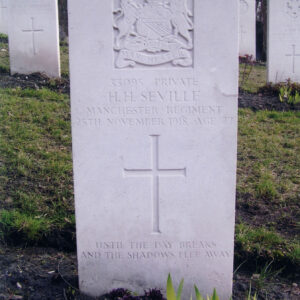 biała, pionowa płyta nagrobna z wyżłobionym krzyżem łacińskim, pośrodku którego wyryto znak Pułku Manchster, za nią inne nagrobki i drzewa