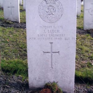 pionowa, biała płyta nagrobna z wyżłobionym krzyżem łacińskim, inskrypcją i znakiem Pułku Saperów, za nią inne nagrobki i drzewa