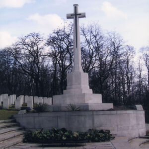 Wysoki krzyż z aplikacją miecza z brązu na cokole pośrodku kolistej platformy z zielenią. Po lewej stronie schody prowadzące do nagrobków. Z tyłu drzewa bez liści.