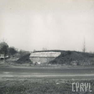 skrzyżowanie dwóch ulic, obok których stoi usytuowana w nasypie ziemnym budowla z cegieł; widoczne drzewa oraz jadące samochody