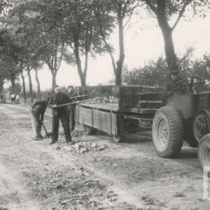 grupa mężczyzn w strojach roboczych z łopatami, pracująca na piaszczystej, nieutwardzonej drodze, dwóch z nich ładuje łopatami kamienie na przyczepę przypięta do ciągnika, na którym siedzi mężczyzna; po obu stronach drogi rosną drzewa