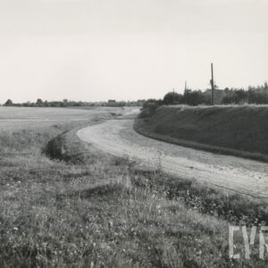 polna droga, po jej jednej stronie rozpościerają się pola, po drugiej wał ziemny, a na nim drzewa i wolnostojące domy mieszkalne, w oddali drzewa i domy