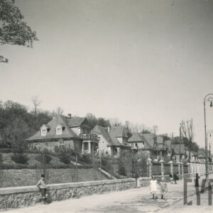 brukowana droga z chodnikiem, po którym spacerują dzieci, wzdłuż ulicy stoją lampy i słupy energetyczne, głębiej wille mieszkalne