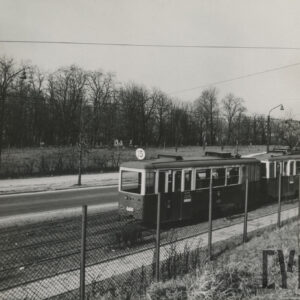 ulica z jadącym tramwajem, w głębi drzewa, nagrobki i budynek mieszkalny