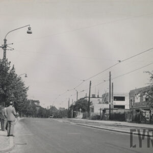 ulica skręcająca w prawo równolegle do torów tramwajowych, po jej lewej stronie drzewa i lampy uliczne i przystanek autobusowy, po drugiej stronie budynki mieszkalne