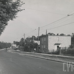 ulica skręcająca w prawo równolegle do torów tramwajowych, po których jedzie tramwaj; po jej lewej stronie drogi widoczne drzewa i lampy uliczne, po prawej stronie budynki mieszkalne i słupy energetyczne