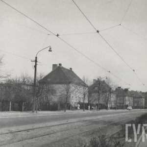 ulica skręcająca z biegnącymi równolegle torami tramwajowymi; po lewej stronie drogi widoczni ludzie spacerujący chodnikiem, za nimi żywopłoty i wolnostojące domy mieszkalne