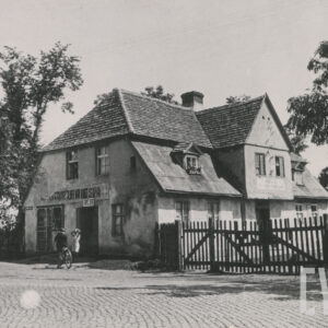 murowany dom z cegieł, pokryty dachówką, otoczony płotem; budynek stoi utwardzanej, brukowanej drodze, wzdłuż której rosną drzewa i stoją inne budynki mieszkalne