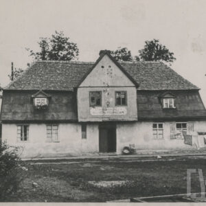 murowany dom z cegieł ze zniszczoną elewacją, pokryty dachówką, otoczony płotem; na sznurkach wisi pranie