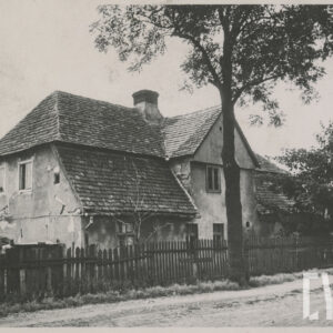 murowany dom z cegieł, pokryty dachówką, otoczony płotem; budynek stoi przy nieutwardzanej, piaszczystej drodze