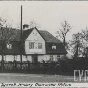 murowany dom z cegieł, pokryty dachówką, otoczony płotem i drzewami; budynek stoi przy utwardzanej, brukowanej drodze