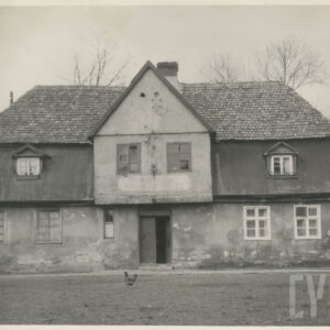 murowany dom z cegieł, pokryty dachówką, otoczony płotem; przed budynkiem widoczne piaszczyste podwórze