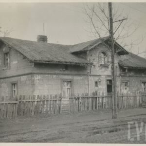 niski dom murowany, otoczony drewnianym płotem, stojący przy brukowanej drodze