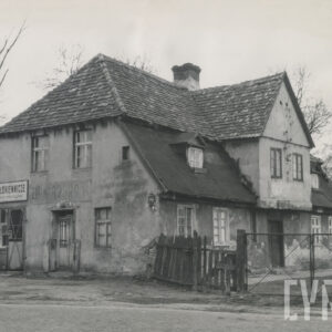 murowany dom z cegieł, pokryty dachówką, otoczony płotem; budynek stoi przy utwardzanej, brukowanej drodze