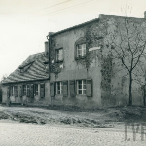 niski murowany budynek mieszkalny z cegieł z dobudówką; spod zniszczonej warstwy elewacji widoczne gdzieniegdzie cegły; część okien posiada okiennice