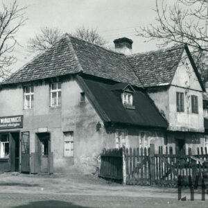 murowany dom z cegieł, pokryty dachówką, otoczony płotem; budynek stoi przy utwardzanej, brukowanej drodze