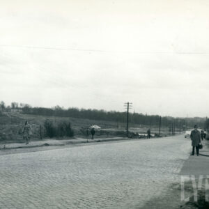 brukowana ulica, wzdłuż której stoją słupy energetyczne, w oddali drzewa; wzdłuż ulicy widać spacerujących ludzi oraz jeżdżące samochody