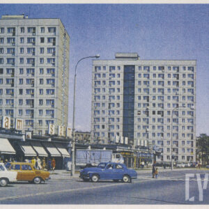 samochody zaparkowane przy pawilonach handlowych, obok ulicy, na której widać przejście dla pieszych, w oddali bloki z wielkiej płyty