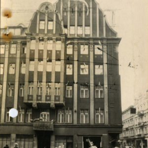 Elewacja budynku hotelu przed przebudową. Liczne okna z flagami, dekoracyjny szczyt. Przed budynkiem idący ludzie w zimowych strojach.