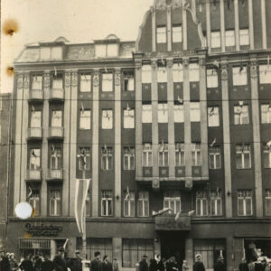 Fasada budynku z ozdobnymi szczytami. Liczne okna z flagami. Przed hotelem idący ludzie w zimowych strojach.