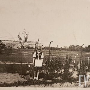 mała dziewczynka w stroju krakowskim stoi w ogrodzie, za nią druciany płot i rozległe nieużytki, z prawej fragment domu na narożniku ulic Kasprzaka i Hetmańskiej