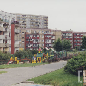 na pierwszym planie plac zabaw dla dzieci z huśtawkami, małymi kolorowymi domkami, w głębi pięciokondygnacyjne bloki mieszkalne, za nimi wysoki blok
