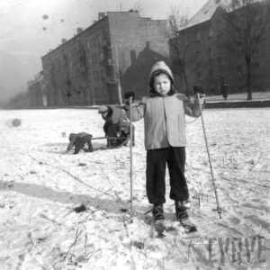 dziewczynka na nartach trzyma w rękach kijki, w głębi bloki mieszkalne, wokół śnieg