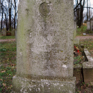 zabrudzona i pokryta mchem, kamienna płyta ze słabo czytelnymi napisami i śladem po medalionie portretowym, za nią inne nagrobki i drzewa bez liści