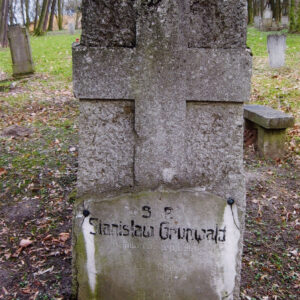 górna część płyty nagrobnej o chropowatej fakturze z wykutym krzyżem łacińskim, w dolnej części zabrudzona tablica z inskrypcją, z tyłu teren cmentarza z innymi nagrobkami i drzewami