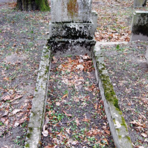 zniszczona płyta nagrobna z odłamanymi górnymi narożami i nieczytelnymi napisami, omszałe obmurowanie mogiły ziemnej, wokół teren cmentarza i fragmenty innych nagrobków