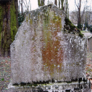 zabrudzona kamienna płyta z odłamanymi górnymi narożami i nieczytelnymi napisami, za nią teren cmentarza, fragmenty nagrobków i pnie drzew