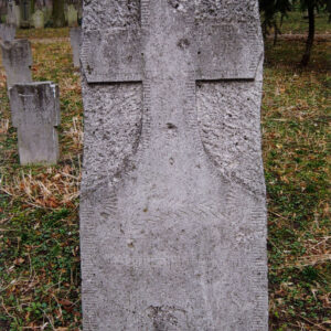 płyta nagrobna o chropowatej powierzchni z wykutym krzyżem łacińskim, w dolnej części zatarta inskrypcja, z tyłu teren cmentarza, inne nagrobki i drzewa 