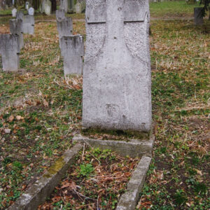 obmurowana mogiła ziemna i prostokątna stela z wykutym krzyżem i nieczytelnymi napisami, wokół teren cmentarza, inne nagrobki i drzewa