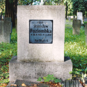 kamienna płyta w kształcie kwadratu na cokole ze szklaną tablicą, na której umieszczono czarne napisy, wokół teren cmentarza