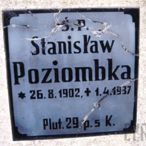 kwadratowa, szklana tablica z napisami w kolorze czarnym i licznymi pęknięciami przytwierdzona do płyty nagrobnej